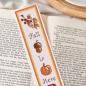 May include: A bookmark with an autumn theme. The bookmark features a cross-stitch design with images of autumn leaves, an acorn, a pumpkin, and a mushroom. The text "Fall is Here" is stitched in the centre of the bookmark.