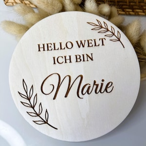 Puede incluir: Un letrero redondo de madera clara con el texto grabado "HELLO WELT ICH BIN Marie" y diseños de hojas. El letrero está rodeado de flores secas y una cesta de mimbre.