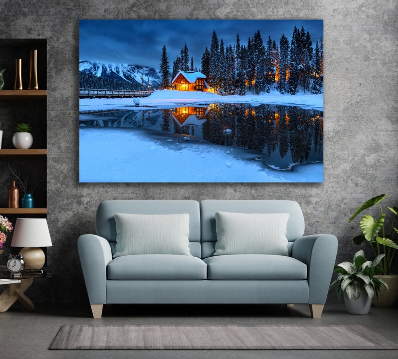 May include: A wooden cabin with lit windows sits on a snow-covered lakeshore, surrounded by a forest of evergreen trees. The cabin is reflected in the still, blue water of the lake.