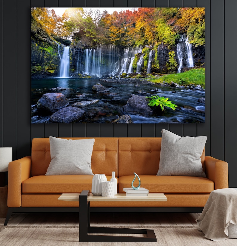 May include: A scenic photograph of a waterfall cascading down a rocky cliff face, surrounded by lush green foliage and vibrant autumn colors. The water flows into a clear pool below, with large rocks scattered throughout the stream.