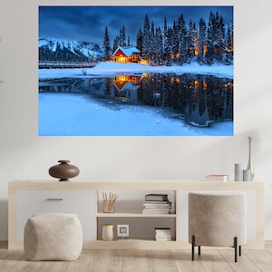 May include: A wooden cabin with lit windows sits on a snow-covered lake surrounded by a forest of pine trees. The lake is frozen and reflects the cabin and trees. The sky is a dark blue with clouds.