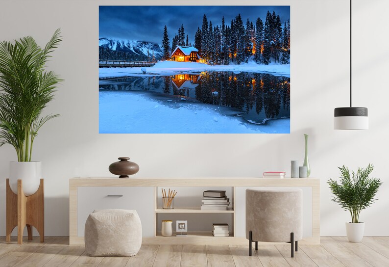 May include: A wooden cabin with lit windows sits on a snow-covered lake surrounded by a forest of pine trees. The lake is frozen and reflects the cabin and trees. The sky is a dark blue with clouds.