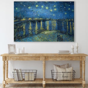 May include: A painting of a starry night sky over a river with a city skyline in the distance. The painting is in a blue and yellow color scheme and features a swirling brushstroke style. The painting is hanging on a white wall above a wooden console table with two baskets and a pillow.