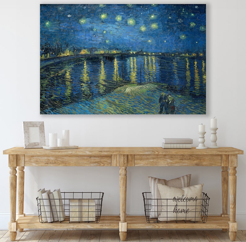 May include: A painting of a starry night sky over a river with a city skyline in the distance. The painting is in a blue and yellow color scheme and features a swirling brushstroke style. The painting is hanging on a white wall above a wooden console table with two baskets and a pillow.