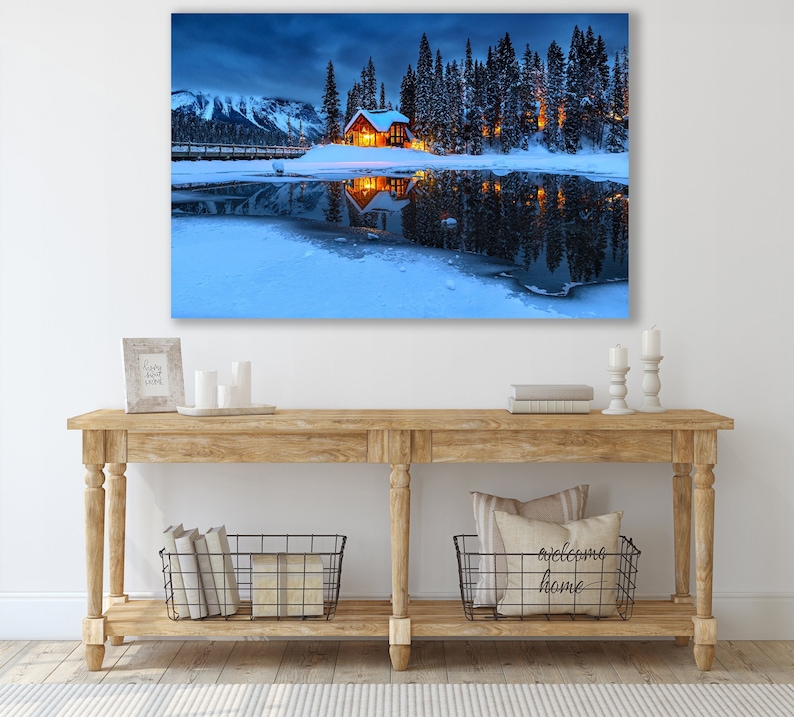 May include: A cozy cabin with glowing windows sits on a snow-covered island in a frozen lake. The cabin is surrounded by tall evergreen trees and the reflection of the cabin and trees can be seen in the water.