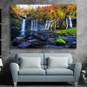 May include: A scenic photograph of a waterfall cascading down a rocky cliff face, surrounded by lush green trees with vibrant autumn foliage. The water flows into a clear pool below, with large rocks scattered throughout the stream.