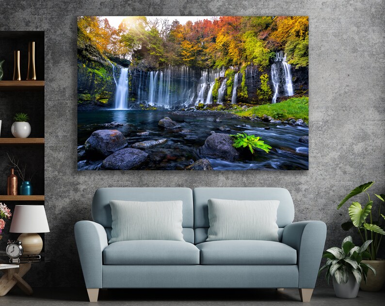 May include: A scenic photograph of a waterfall cascading down a rocky cliff face, surrounded by lush green trees with vibrant autumn foliage. The water flows into a clear pool below, with large rocks scattered throughout the stream.