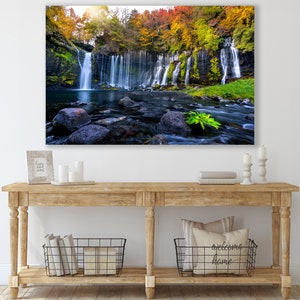 May include: A photograph of a waterfall cascading down a rocky cliff face, surrounded by lush green foliage and vibrant autumn colors. The water flows into a clear pool below, with smooth rocks scattered throughout.