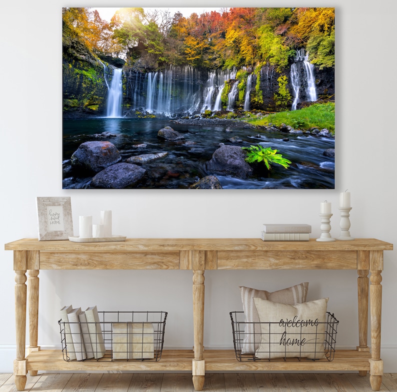 May include: A photograph of a waterfall cascading down a rocky cliff face, surrounded by lush green foliage and vibrant autumn colors. The water flows into a clear pool below, with smooth rocks scattered throughout.