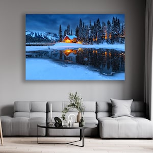 May include: A small wooden cabin with lit windows sits on a snow-covered lake shore, surrounded by tall evergreen trees. The cabin is reflected in the still water, creating a symmetrical image. The mountains in the background are covered in snow and the sky is a deep blue.