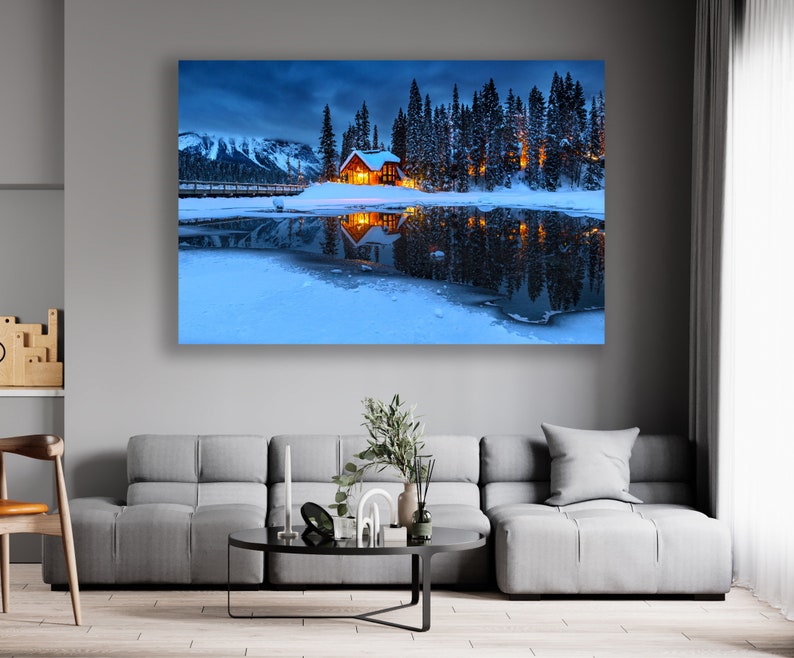 May include: A small wooden cabin with lit windows sits on a snow-covered lake shore, surrounded by tall evergreen trees. The cabin is reflected in the still water, creating a symmetrical image. The mountains in the background are covered in snow and the sky is a deep blue.