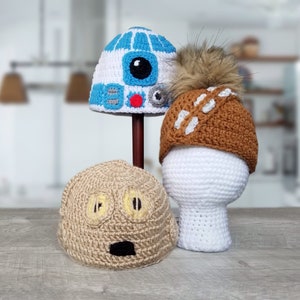 May include: Three crocheted hats, one blue and white with a robot face, one brown with a white X and a brown fur pom pom, and one tan with a cartoon face.