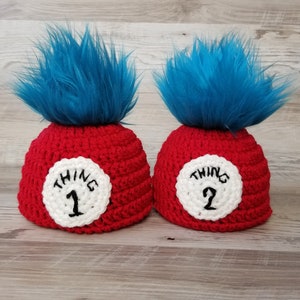Könnte beinhalten: Zwei rote gehäkelte Hüte mit blauem Kunstfell und weißen Kreisen mit dem Text "THING 1" und "THING 2" in Schwarz. Die Hüte sind wahrscheinlich für ein Kostüm oder eine Themenveranstaltung.