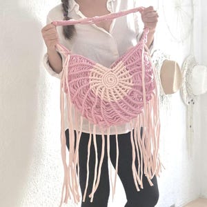 May include: A handmade macrame shoulder bag in shades of pink and cream, featuring a circular design and long fringe. The bag is held by a person wearing a white shirt, black leggings, and beige boots.