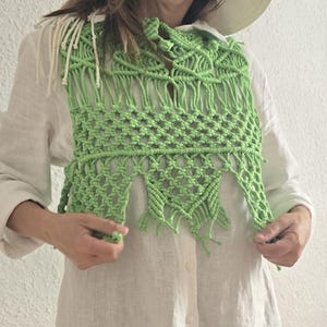 Op de afbeelding: Een lichtgroen macramé vest met franjes. Het vest is gemaakt van geknoopt katoenen koord en heeft een geometrisch patroon. Het wordt gedragen over een wit overhemd. Het macramé vest is een unieke, bohemien stijl.