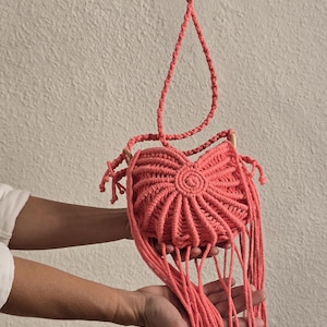 May include: Coral-colored macrame handbag with a circular design and long fringe. The bag has a braided strap and wooden accents. The handmade bag is held up by a person.