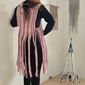 Op de afbeelding: Een persoon draagt een crèmekleurige hoed en een roze en witte gehaakte vest met lange franjes. Ze draagt een zwart shirt met lange mouwen, een zwarte legging en beige laarzen. De achtergrond omvat macramé wandkleden en een decoratieve lamp.