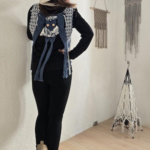 Op de afbeelding: Een handgemaakt macramé vest in blauw en wit, met een uilontwerp en houten kralen. Het vest wordt gedragen over een zwart shirt met lange mouwen, gecombineerd met een zwarte broek en beige laarzen. Macramé wandkleden zijn op de achtergrond te zien.