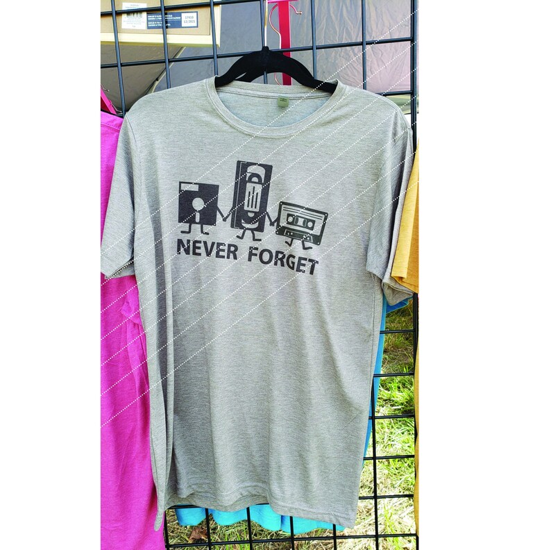 Puede incluir: Una camiseta gris con un gr&aacute;fico de un disquete, una cinta VHS y una cinta de cassette con el texto "NEVER FORGET".
