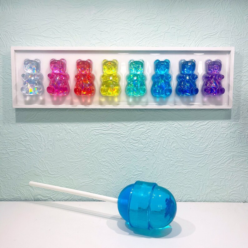 May include: A white frame with eight gummy bear figurines in a rainbow of colors. The gummy bears are arranged in a row and are all facing forward. The gummy bears are made of clear plastic and have a shiny finish. There is a blue lollipop on a white stick in the foreground.
