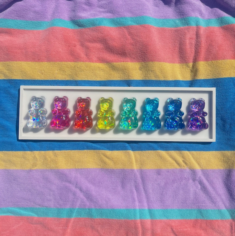 May include: A white tray with eight gummy bear figurines in a rainbow of colors. The gummy bears are clear and iridescent, with a shiny finish. The tray is on a striped fabric background.