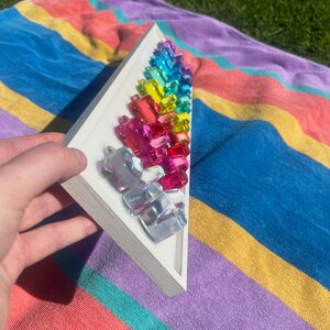 May include: A white frame with a rainbow of colorful glass pieces arranged in a triangular shape. The glass pieces are iridescent and reflect light.