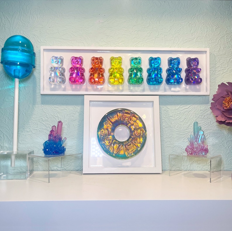 May include: A rainbow assortment of gummy bear figurines displayed on a white shelf with a blue lollipop and two crystal-like decorations.