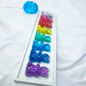 May include: A white frame with a row of gummy bear figurines in rainbow colors, including red, orange, yellow, green, blue, purple, and clear. The gummy bears are arranged in a line on a white background.