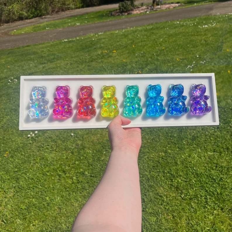 May include: A white display case with eight iridescent gummy bear figurines in a rainbow color gradient. The gummy bears are arranged in a row from left to right, starting with clear, then pink, red, yellow, green, blue, light blue, and purple.