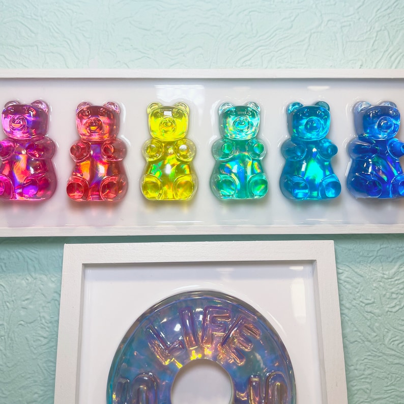 May include: A set of six gummy bear shaped candies in a white frame. The gummy bears are iridescent and come in the colors red, pink, yellow, green, blue, and purple.