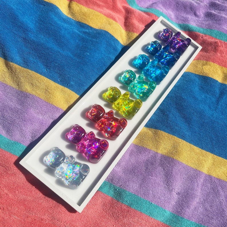 May include: A white tray with a rainbow assortment of gummy bear shaped resin charms. The charms are iridescent and have a holographic finish.