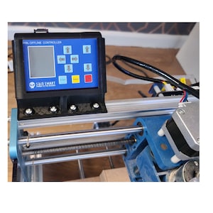 May include: A black and blue CNC machine with a control panel labelled "GRBL OFFLINE CONTROLLER" and a screen displaying arrows. The control panel also has buttons labelled "STEP", "TUNE", and "RUN".