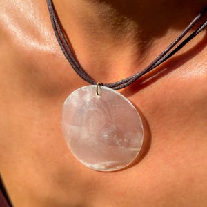 Può includere: Un ciondolo circolare, traslucido e rosa pallido con una superficie liscia e lucida, sospeso a una collana con cordino marrone scuro a più fili. Il ciondolo presenta sottili inclusioni bianche nuvolose e un morbido riflesso sulla superficie.