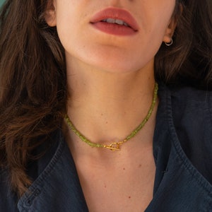 May include: A close-up of a woman wearing a dark blue sleeveless top and a gold necklace with green gemstone beads. The necklace has a gold toggle clasp.