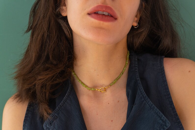 May include: A close-up of a woman wearing a dark blue sleeveless top and a gold necklace with green gemstone beads. The necklace has a gold toggle clasp.
