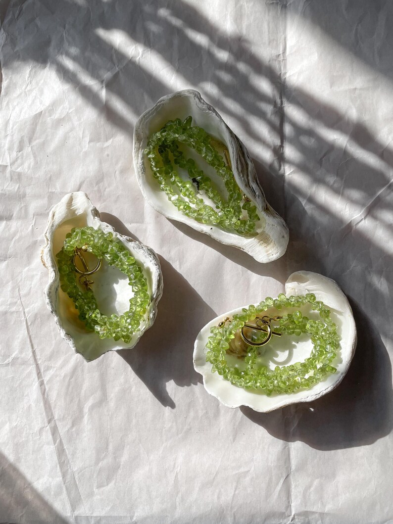 May include: Three white seashells with green gemstone necklaces inside. The necklaces are made of small, irregular shaped green stones and have gold clasps.