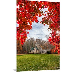 May include: A stone castle with a green roof is framed by vibrant red autumn leaves. The castle is surrounded by a grassy lawn and a forest of trees.