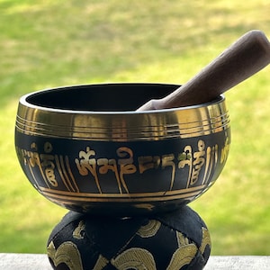 May include: A black and gold singing bowl with a wooden mallet, on a black cushion with gold embroidery. The bowl features gold lettering. The background is a blurred green, suggesting a garden setting.