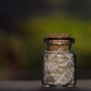 Könnte beinhalten: Eine kleine Glasflasche mit einem Korkverschluss, gefüllt mit weißem, durchscheinendem, papierähnlichem Material.