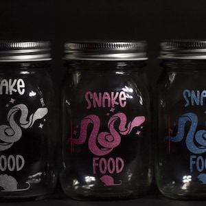 Peut inclure: Trois bocaux en verre avec des couvercles argentés, chacun décoré d'un motif de serpent et des mots "Snake Food". Les serpents sont de couleurs différentes : argent, rose et bleu.
