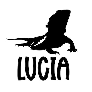 May include: A black silhouette of a bearded dragon lizard with the name "LUCIA" in bold, black letters below. The lizard is depicted in a side profile, with its tail extended and legs visible.
