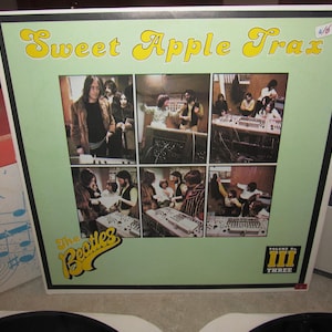 Op de afbeelding: Een vintage albumhoes voor "Sweet Apple Trax" van The Beatles. De hoes heeft een lichtgroene achtergrond met zes kleine foto's van de band in een opnamestudio. De albumtitel is geel en het logo van de band staat linksonder.