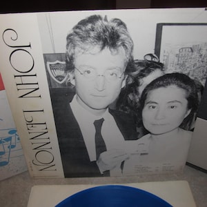 May include: A monochrome album cover featuring John Lennon and Yoko Ono. The album cover has the name "JOHN LENNON" printed vertically. The image shows a close-up of the couple, with Lennon wearing spectacles and a suit and tie.