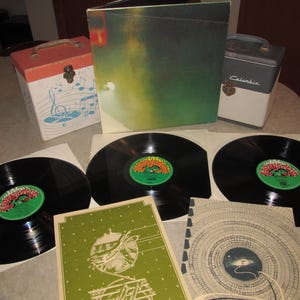 May include: A collection of vintage vinyl records, album covers, and storage cases. The records are black with colorful labels. The album covers feature green and white designs. A red and white music-themed storage box is also present.