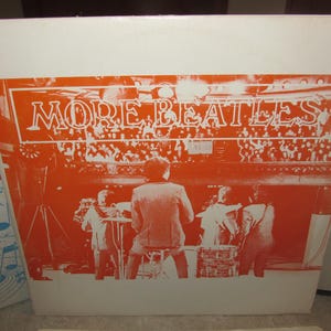 Puede incluir: Una portada de disco de vinilo blanco con fondo naranja. La portada del disco presenta el texto "MORE BEATLES" en letras blancas. La portada del disco también presenta una foto en blanco y negro de una banda tocando en un escenario.