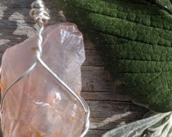 May include: A pink crystal pendant wrapped in silver wire, hanging from a silver chain. The crystal is a raw, natural shape with a rough texture. The pendant is photographed on a wooden surface with green leaves.