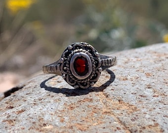 May include: Silver-toned ring with an oval, red gemstone center. The ring features an ornate, detailed design around the gemstone and along the band. The ring is displayed on a textured, gray stone surface.
