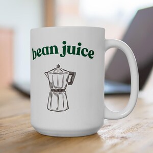 Op de afbeelding: Witte keramische koffiemok met de woorden "bean juice" in groene letters boven een zwart-wit illustratie van een espressomaker. De mok staat op een houten oppervlak.