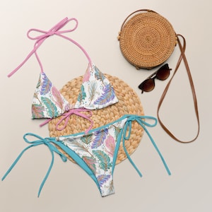 May include: A white bikini set with a colorful feather pattern, featuring pink and turquoise accents. The bikini top has pink ties, and the bottoms have turquoise ties. A round woven bag, sunglasses, and a straw mat complete the beach-themed ensemble.