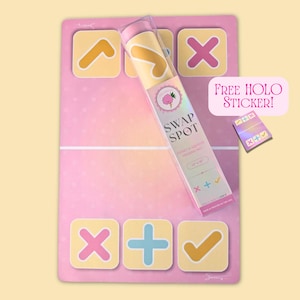 Puede incluir: Una alfombrilla de intercambio rectangular rosa con cuadrados amarillos que contienen símbolos naranjas, rosas y azules. Un envase de tubo de plástico transparente etiquetado Swap Spot Fidget & Squishy Trading Mat 30,48 cm x 45,72 cm descansa encima. Se incluye una pequeña pegatina holográfica.